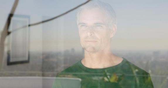 Man looking out of his apartment window, city in reflection | Tom Merton/OJO Images/Getty Images