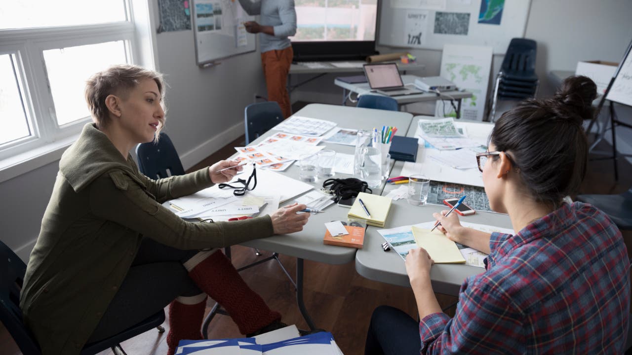 Two women sitting at a cluttered office table working