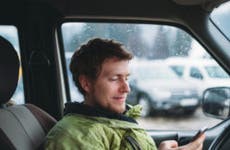 A young man in a green jacket sits in his car, staring down at his phone.